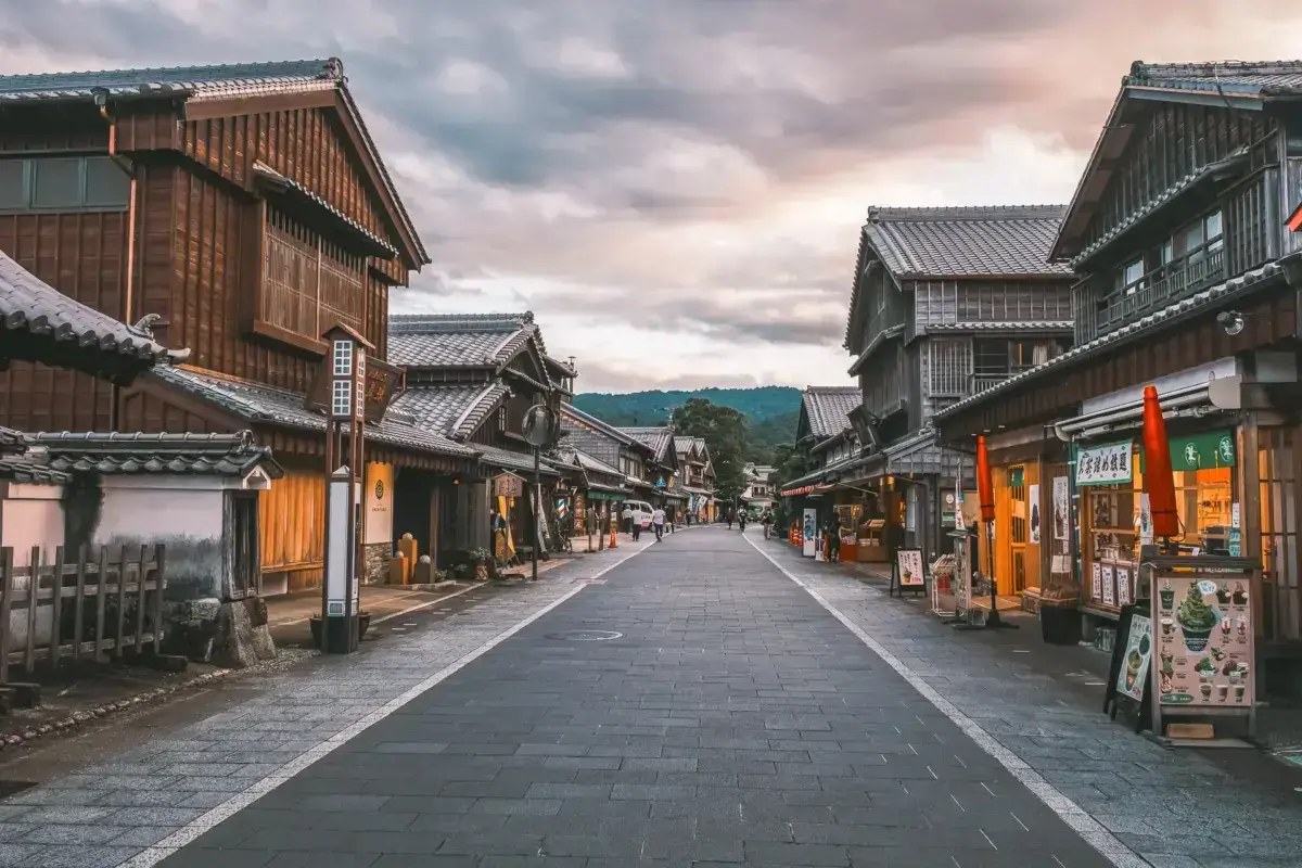 Traditional Japanese street lined with wooden buildings featuring tiled roofs and glowing storefronts, leading toward distant mountains under a dramatic cloudy sky.