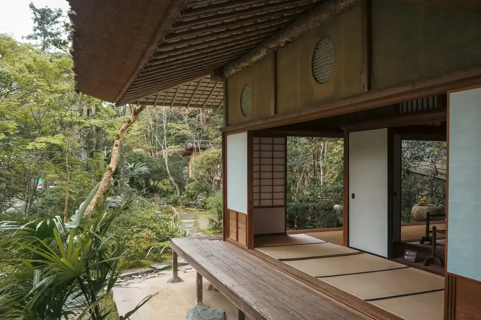 Traditional Japanese house with thatched roof and sliding shoji doors opening onto a wooden veranda, surrounded by a lush garden with green trees and stone pathways.