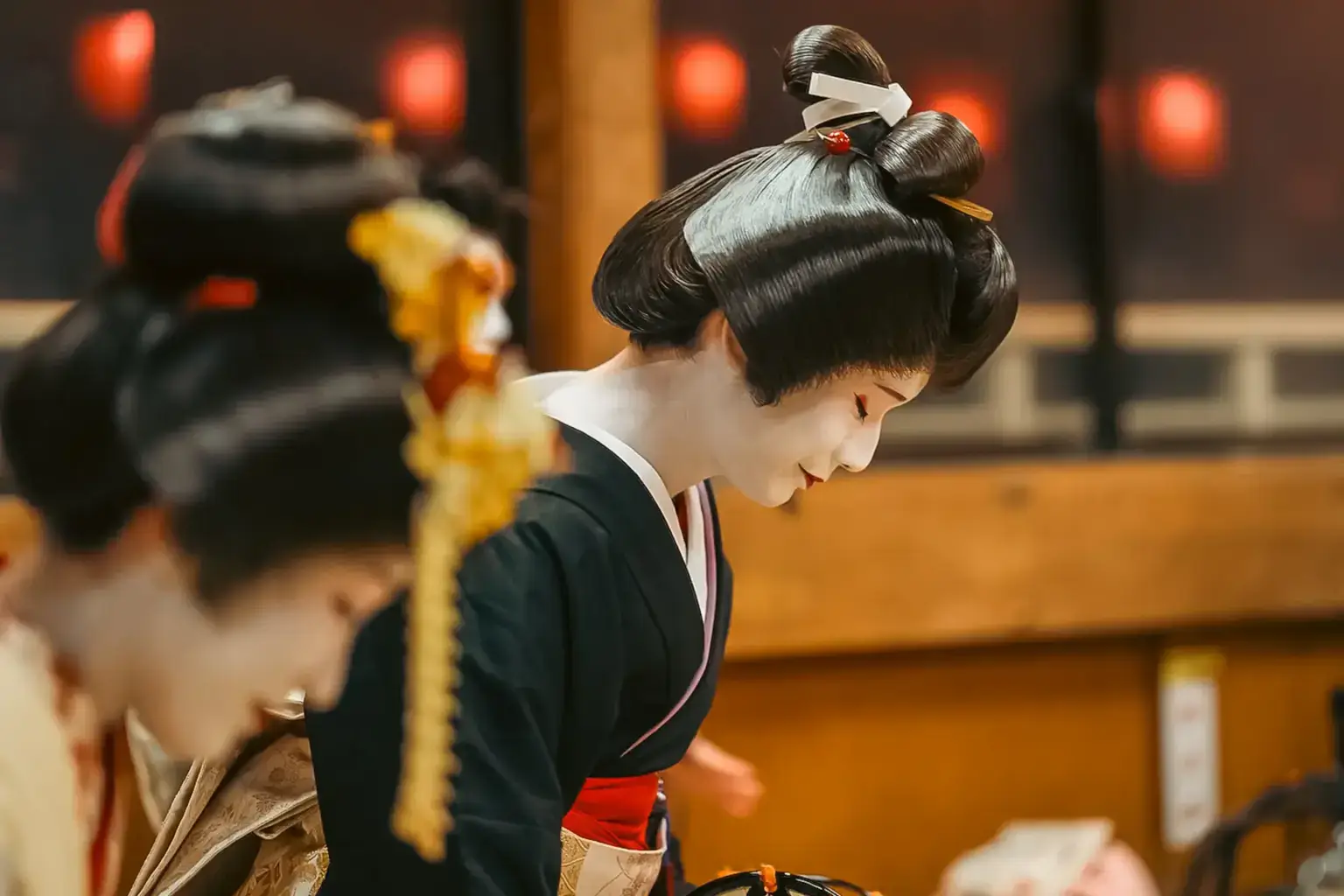 Japanese geisha with traditional shimada hairstyle and dark kimono performing at a temple or traditional venue, with red lanterns glowing in the background.