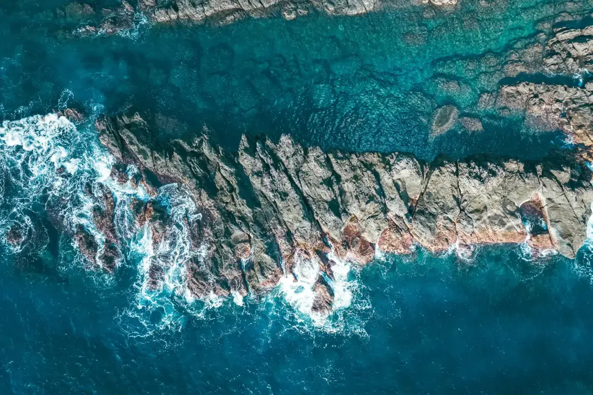 Aerial view of turquoise ocean water flowing around dark jagged coastal rocks with white foaming waves breaking against the rugged shoreline.