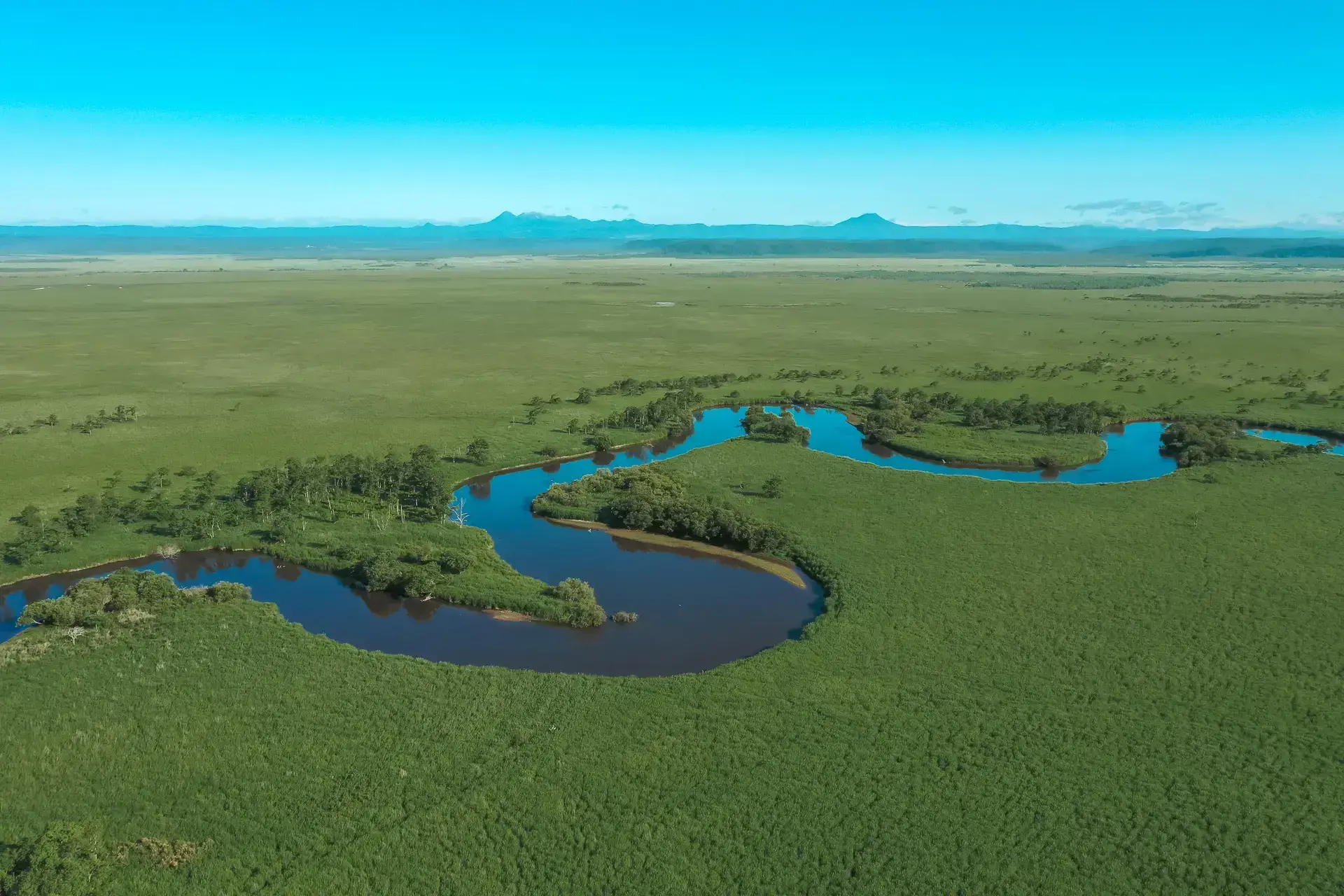 Aerial view of a winding blue river meandering through vast green wetlands and grasslands with distant mountains under a clear sky in East Hokkaido's wilderness.