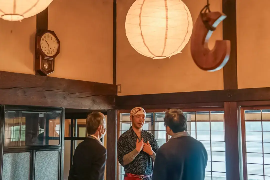 Three people conversing in a traditional Japanese interior with wooden beams, paper lanterns, and shoji screen doors, one person wearing a patterned yukata and headband.
