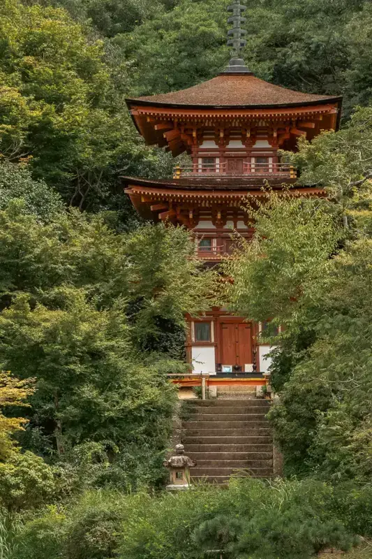 Traditional three-tiered Japanese pagoda with red-brown wooden architecture surrounded by lush green forest, accessed by stone stairs leading to the entrance.