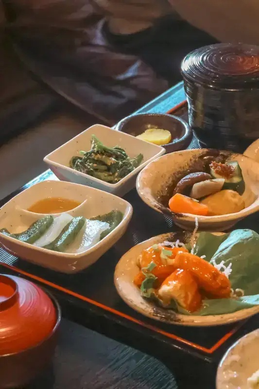 Traditional Japanese set meal featuring golden-battered shrimp tempura, assorted simmered vegetables including pumpkin and mushrooms, sesame-seasoned greens, pickled vegetables, and rice served in individual dishes on a black lacquered tray.