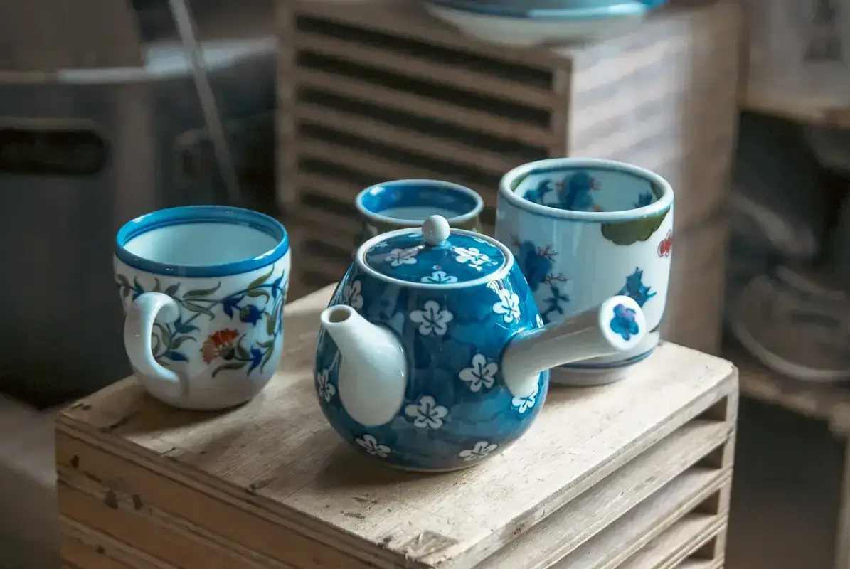 Traditional Japanese tea set with blue and white floral ceramic teapot and two matching cups displayed on a wooden crate in a pottery workshop.
