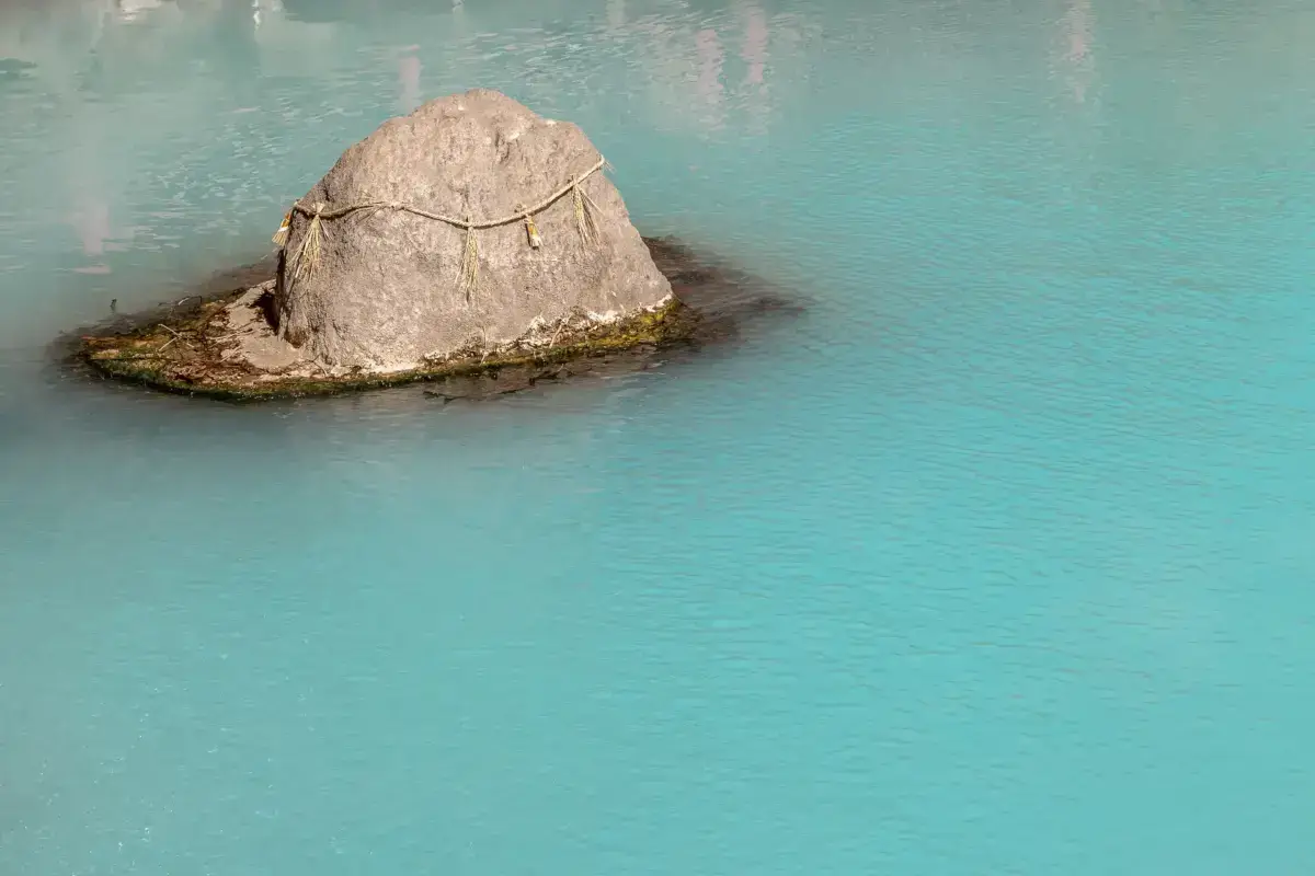 Sacred rock adorned with shimenawa rope sitting in tranquil turquoise hot spring waters in Kyushu, Japan.