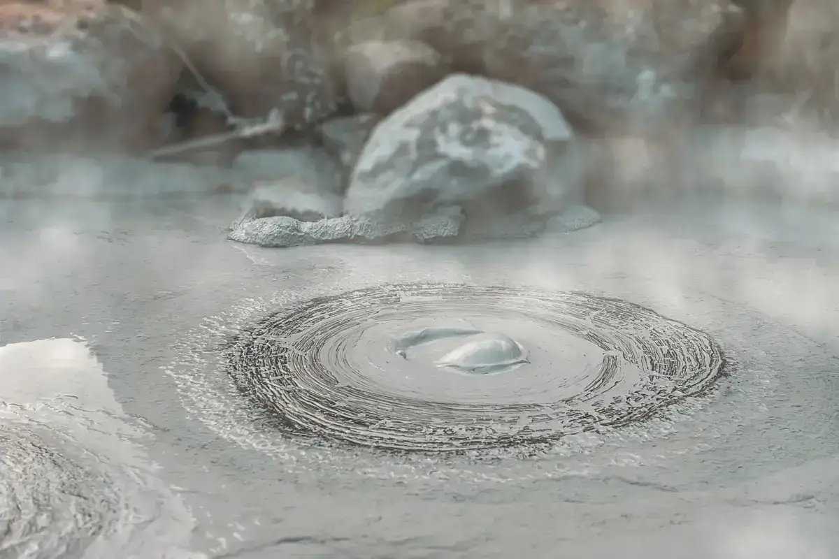 Bubbling mud pool at a geothermal hot spring in Kyushu, with gray volcanic mud creating concentric ripples and steam rising from the heated ground.