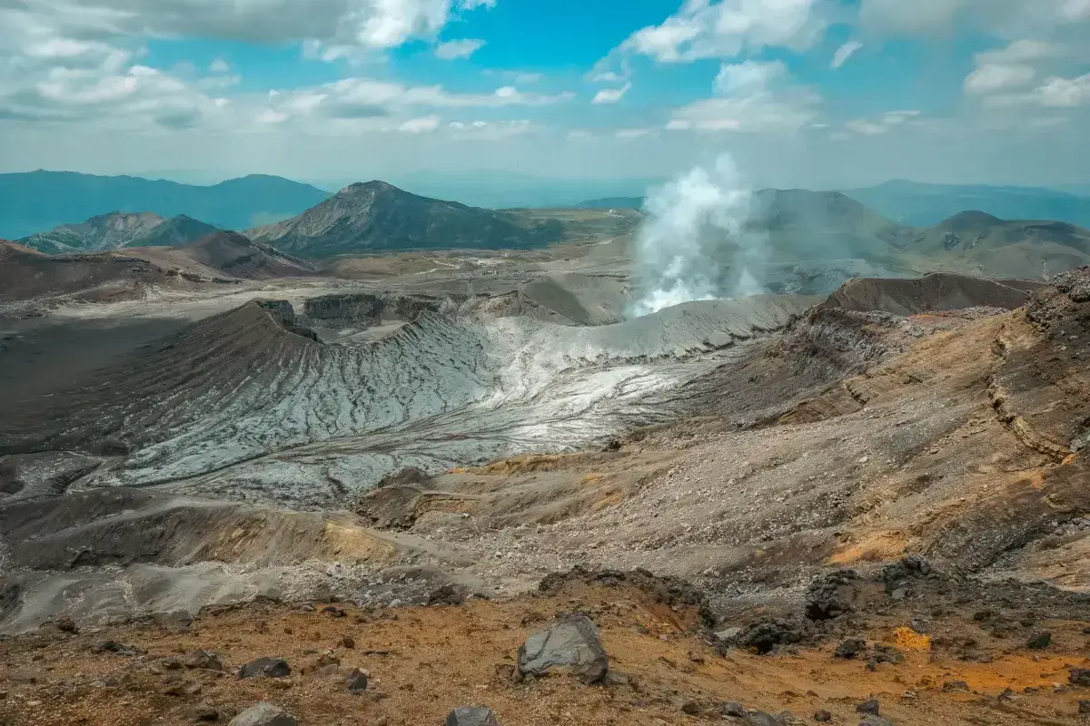 Volcanic landscape in Kyushu showing active steam vents and craters with colorful mineral deposits against a backdrop of mountain ranges under a blue sky.