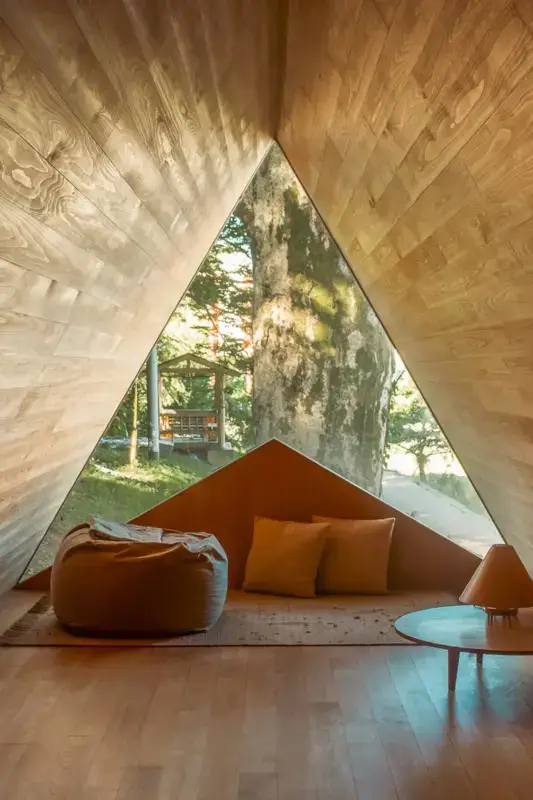 Cozy A-frame reading nook with herringbone wood paneling, featuring a triangular window overlooking forest greenery, warm orange cushions, and a bean bag chair.