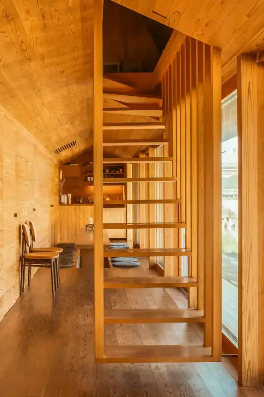 Minimalist wooden staircase with open vertical slat railing in a warm-toned timber interior featuring exposed wood grain walls, ceiling, and flooring with a small dining area visible in the background.