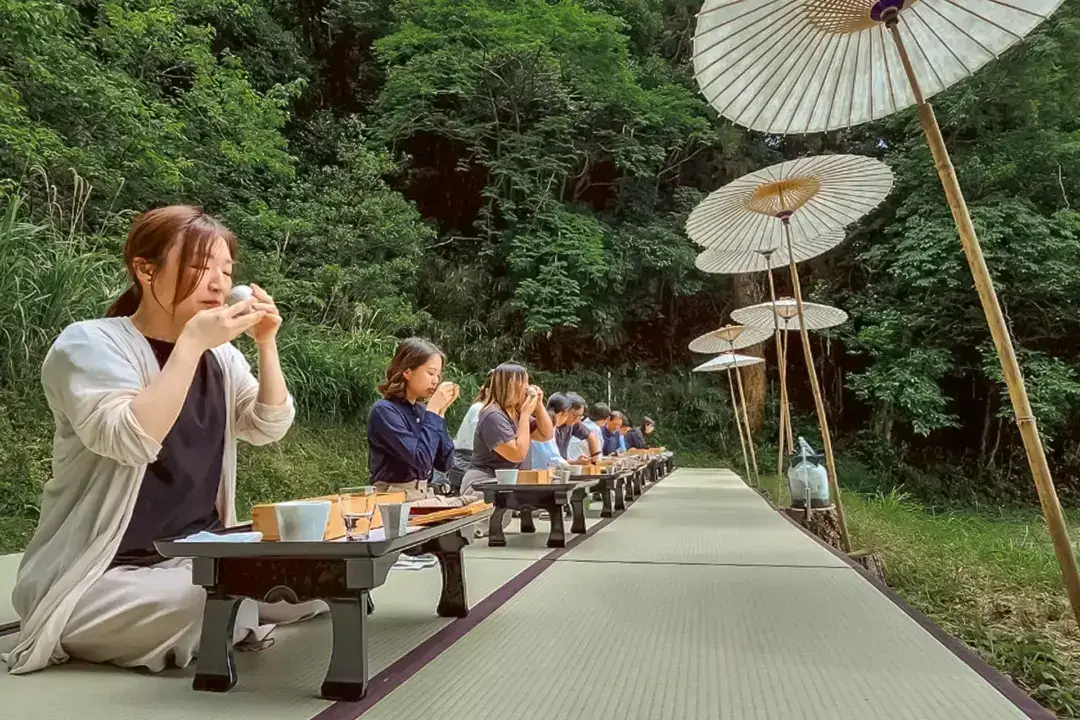 Diners enjoying traditional Japanese multi-course meal on low wooden tables along tatami mat path outdoors, surrounded by lush green forest and shaded by traditional white Japanese parasols.