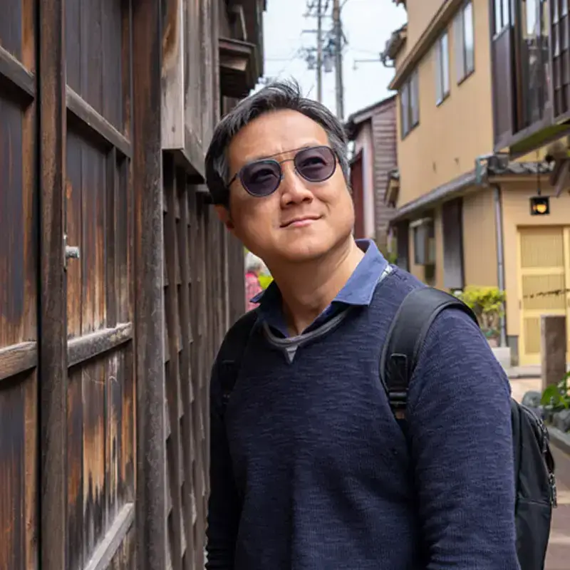 Benjamin Wong wearing sunglasses and a navy sweater, standing on a narrow street in a traditional Japanese neighborhood with wooden buildings.