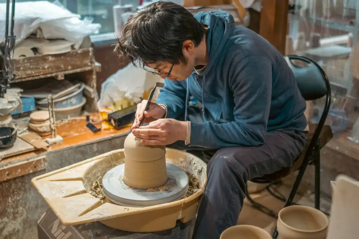 Skilled craftsman in blue shirt carefully shaping a cylindrical clay vessel on a pottery wheel in a workshop, using a carving tool for detailed work.