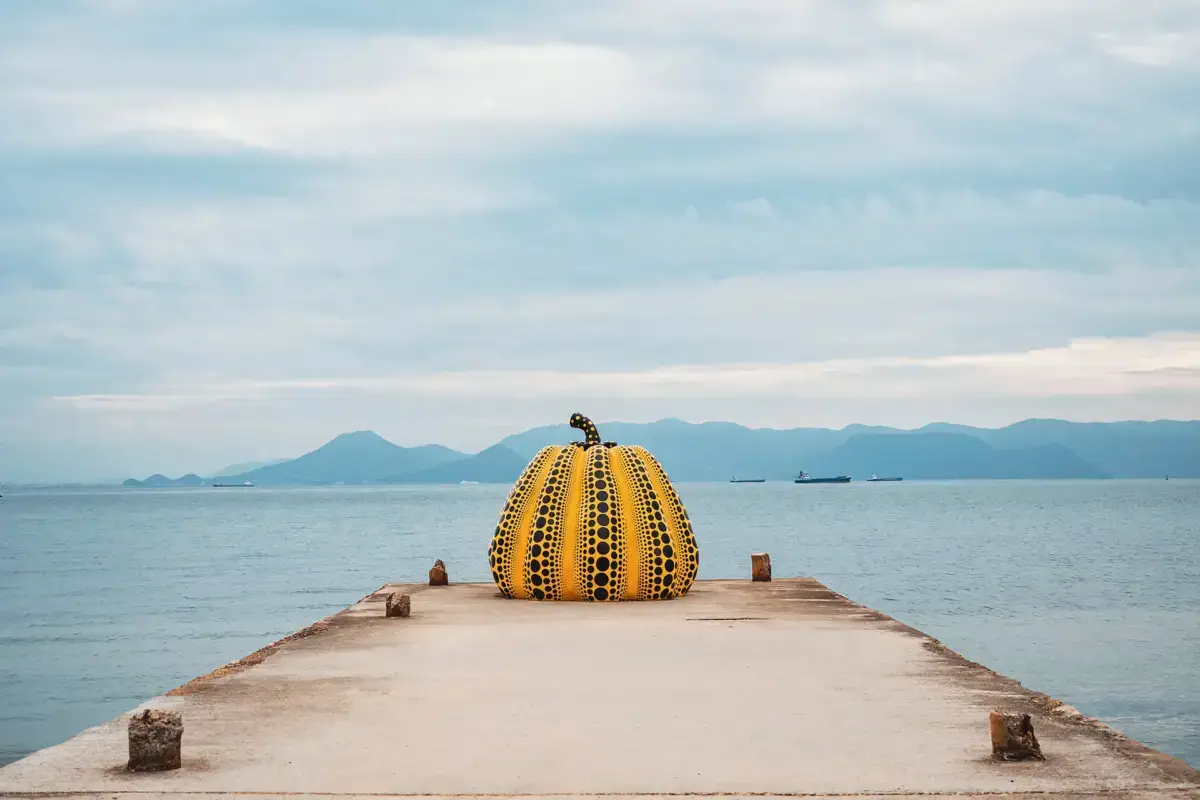 Yellow pumpkin sculpture with black polka dots by Yayoi Kusama on a concrete pier overlooking the Seto Inland Sea with distant mountains under a cloudy sky.