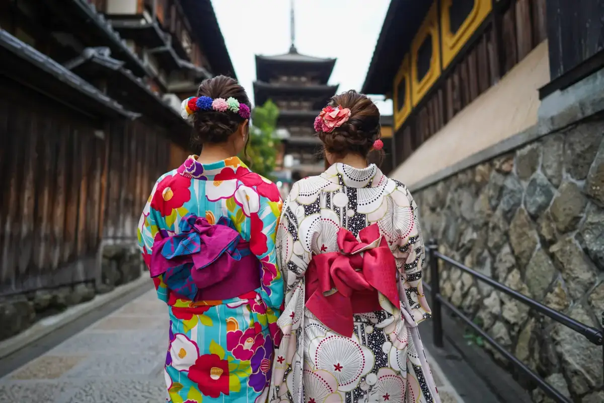 Two women in colorful traditional Japanese kimono with floral hair ornaments walk along a historic street toward a multi-tiered pagoda temple in Kyoto.