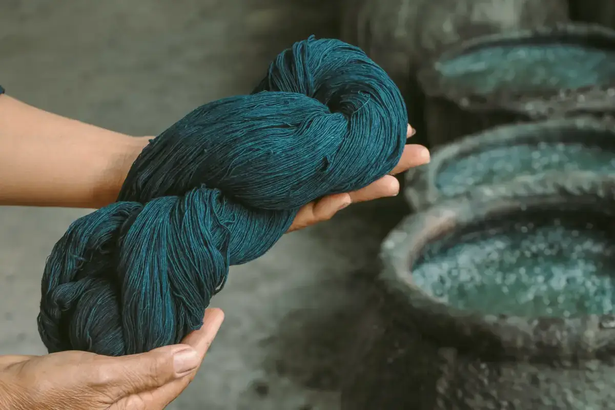 Hands holding a skein of deep teal indigo-dyed yarn above traditional Japanese dyeing vats, showcasing traditional textile craftsmanship.