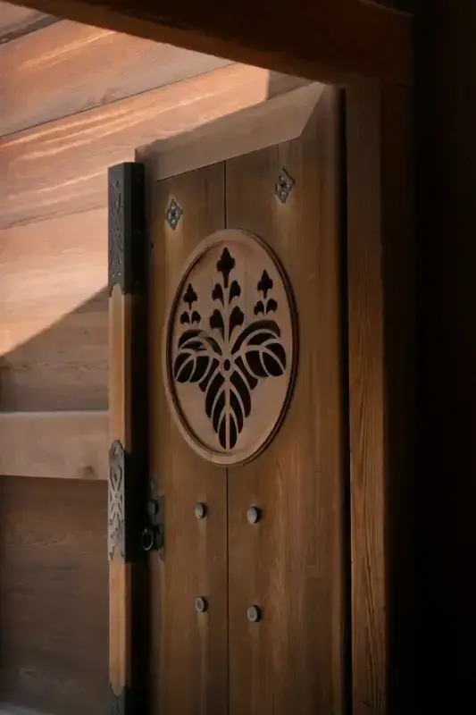Traditional Japanese wooden door featuring an ornamental oval family crest (kamon) with a stylized wisteria motif and decorative metal hardware.
