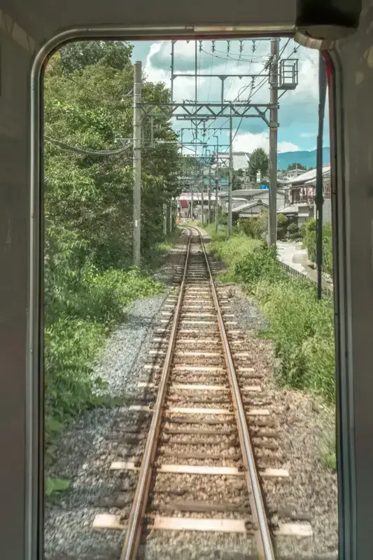 View of railway tracks stretching through a lush green countryside from a train window, with overhead electrical lines, residential buildings, and mountains visible under a blue sky in rural Japan.