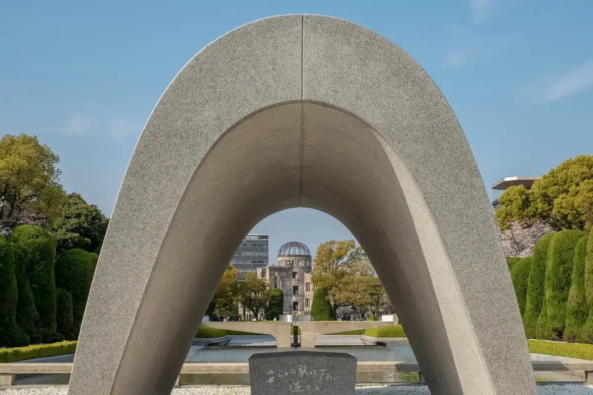 The Hiroshima Peace Memorial Cenotaph with its distinctive concrete arch framing the Atomic Bomb Dome in the background, surrounded by green trees and reflecting pools in Peace Memorial Park.