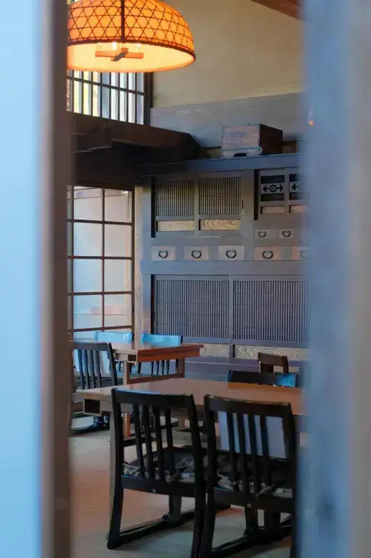 Traditional Japanese dining room with wooden tables and chairs, featuring a woven bamboo pendant light, sliding shoji screens, and built-in storage cabinets with slatted doors.